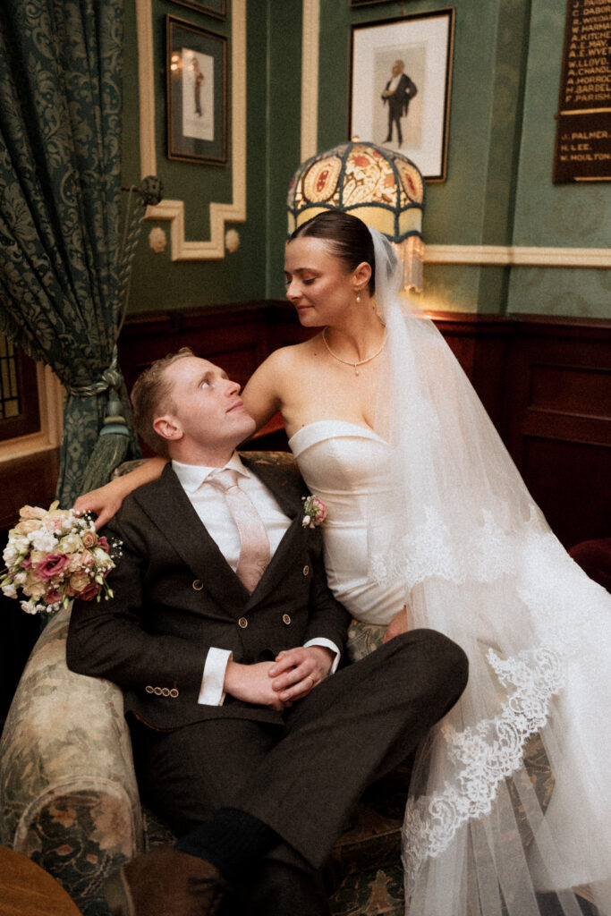 Bride smiling down at the groom seated in an upholstered armchair