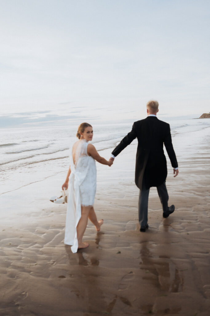 Bride looking back as the couple walk across wet sand