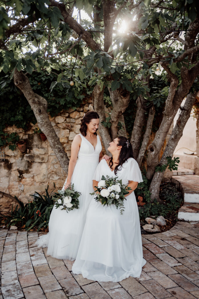 Newlyweds smiling beneath a tree with sunlight filtering through its leaves