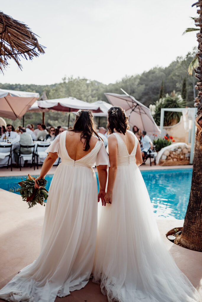Two brides holding hands beside the pool, photographed from behind
