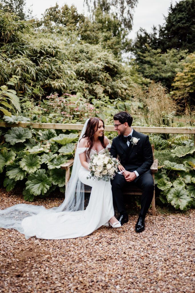 Newlyweds smiling at each other on a bench surrounded by greenery