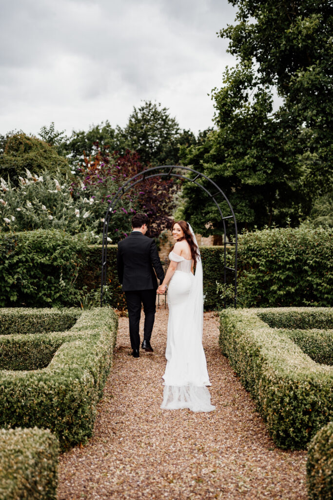 Bride looking back while holding the groom's hand on a garden path