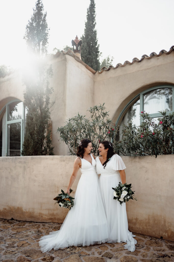 Newlyweds holding bouquets in front of a sunlit courtyard wall