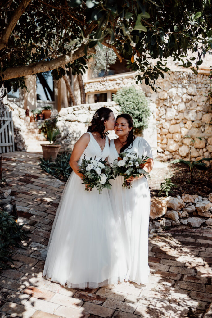 Two brides holding bouquets in dappled shade beside a stone wall