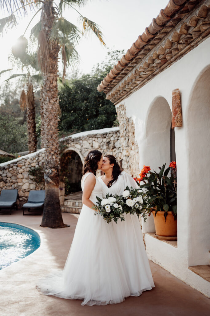 Two brides kissing beside the villa's white arches and swimming pool