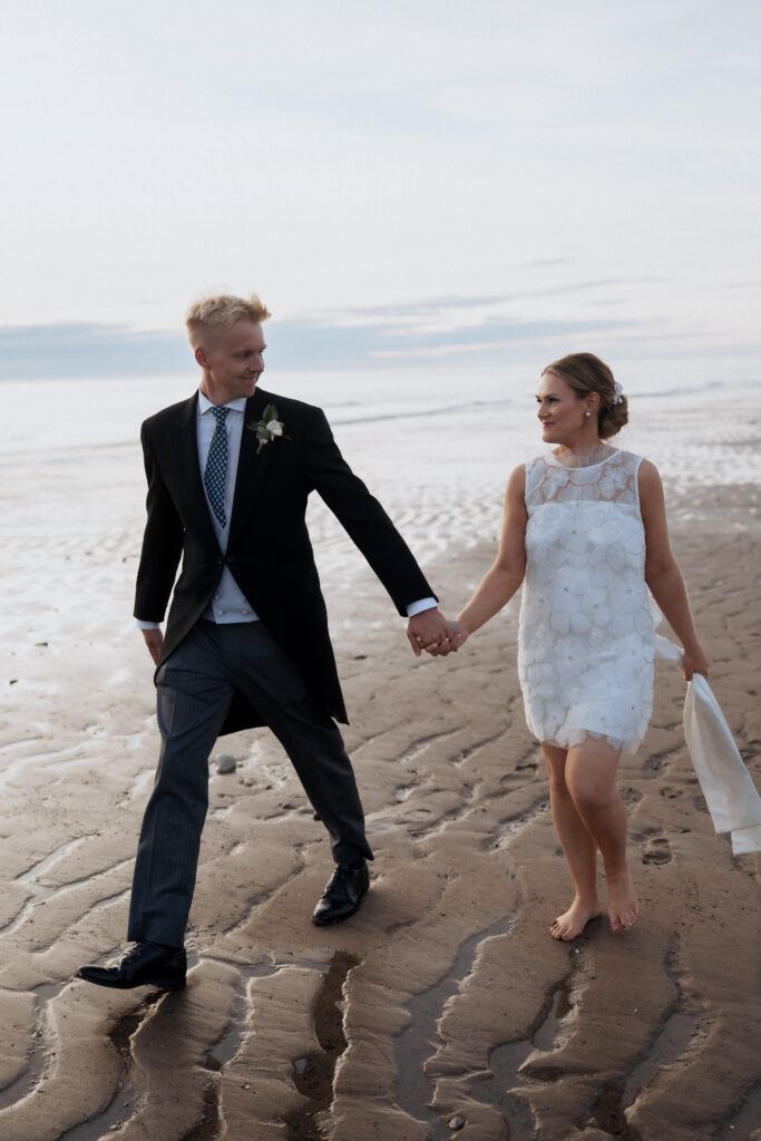 Newlyweds smiling at each other as they walk barefoot along the beach