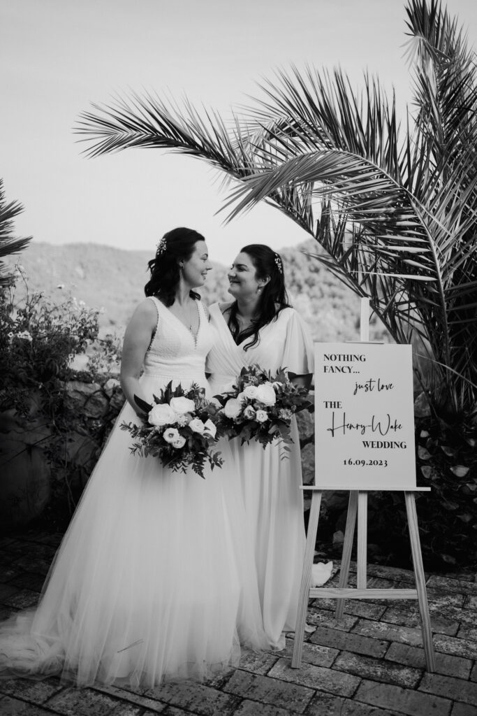 Black-and-white portrait of two brides holding bouquets beside a palm tree