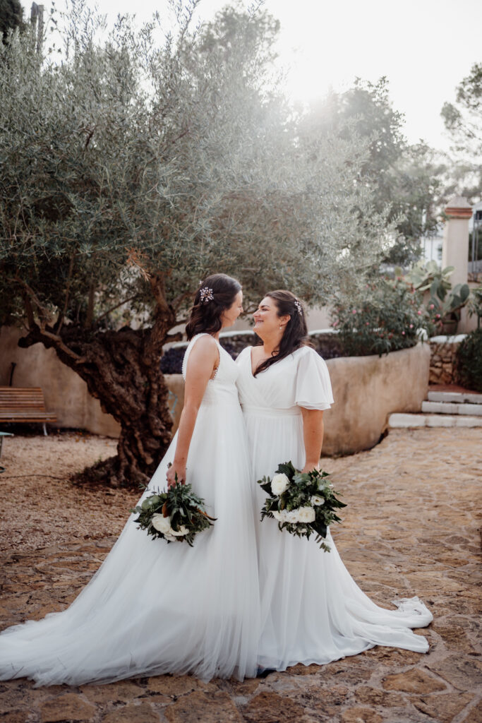 Two brides facing each other beneath a sunlit olive tree