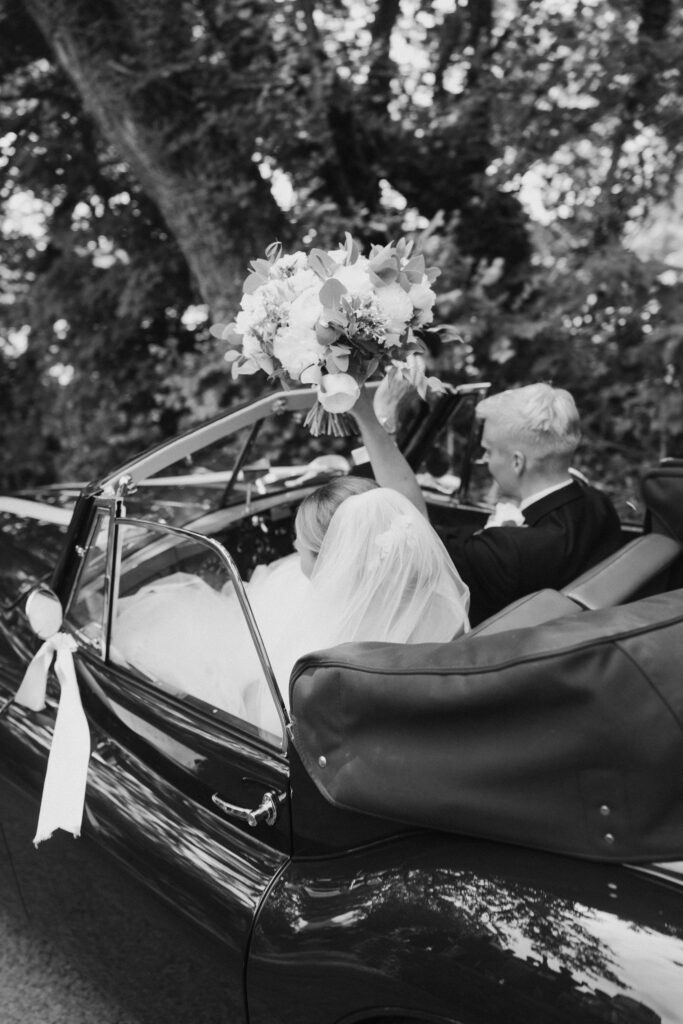 Couple drive off in classic car, bride holding bouquet in the air