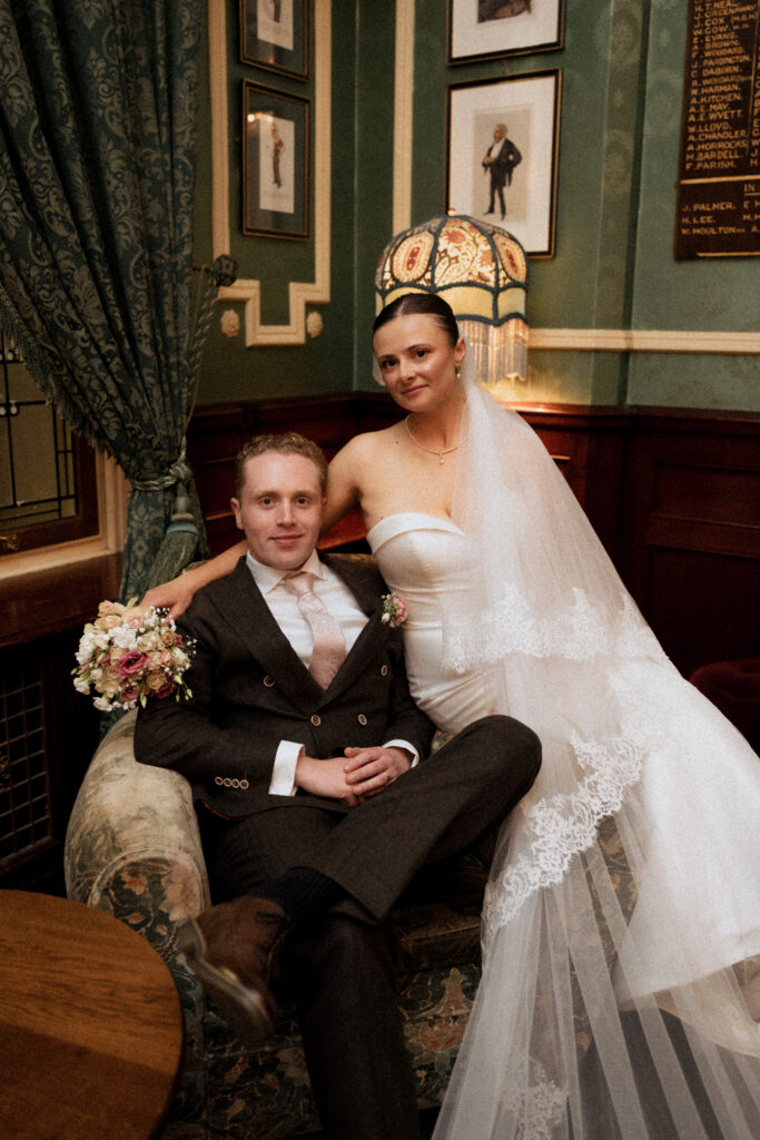 Newlyweds looking towards the camera beside a patterned armchair
