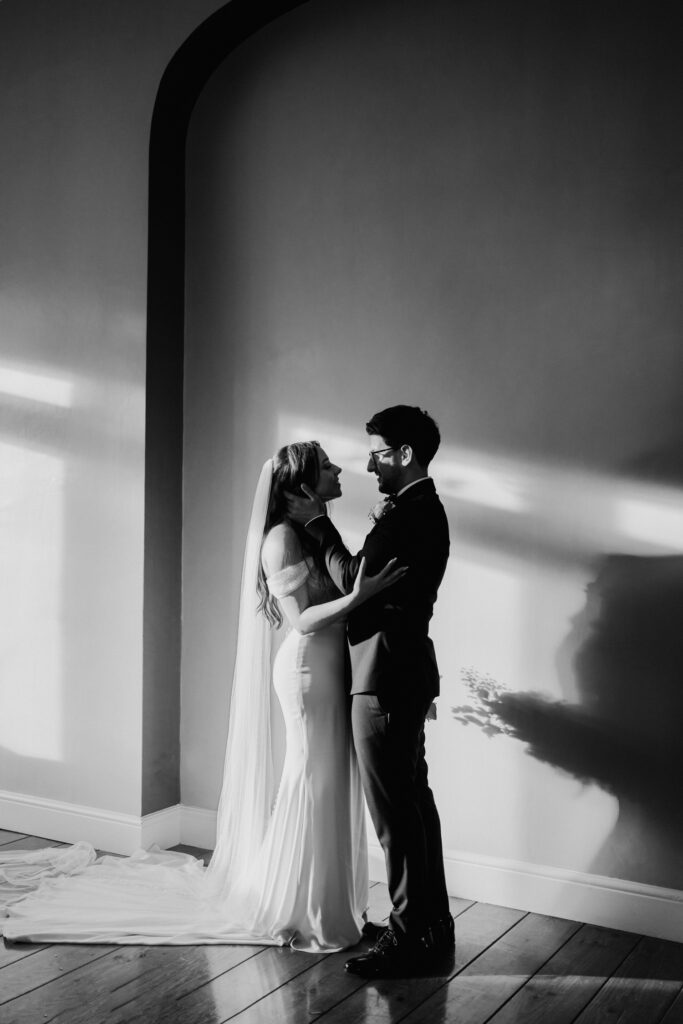 Black-and-white portrait of a couple standing in light from a tall window