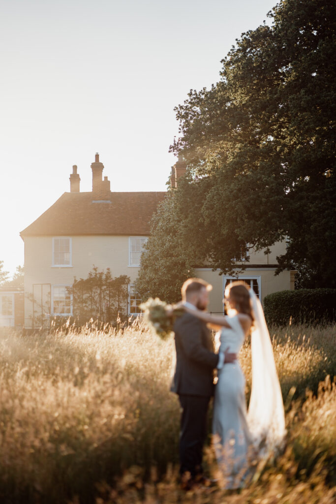 Bride and groom embracing in golden grass beside the house