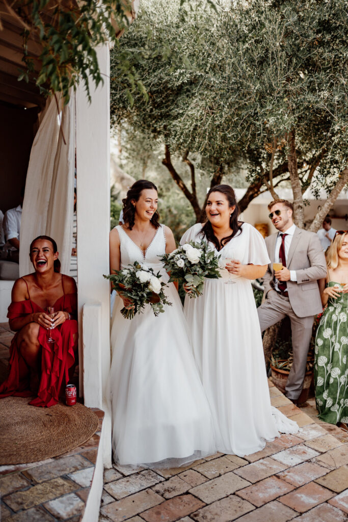 Two brides smiling with bouquets beside the villa doorway