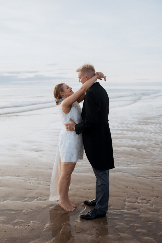 Bride in a short lace dress embracing the groom on the beach