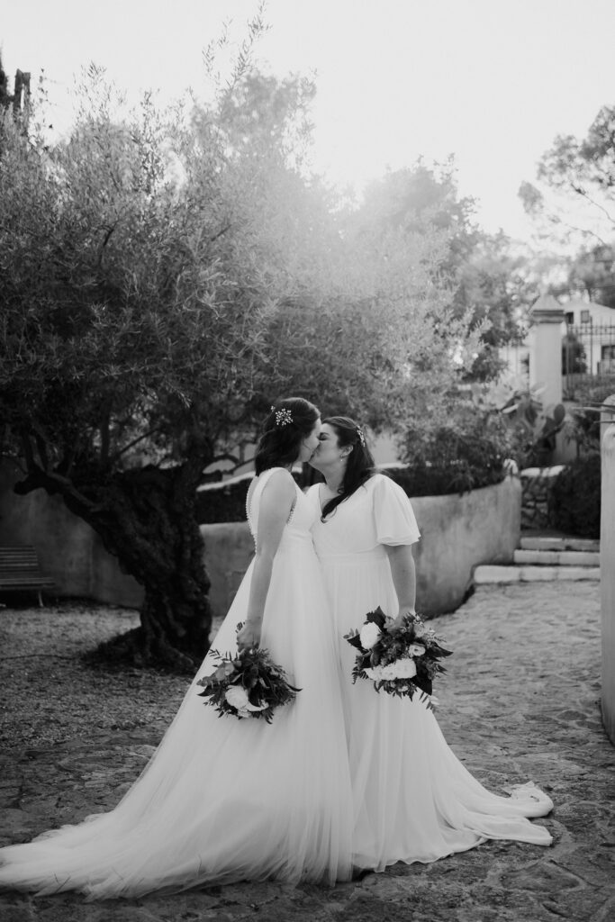 Black-and-white portrait of two brides kissing beside an olive tree