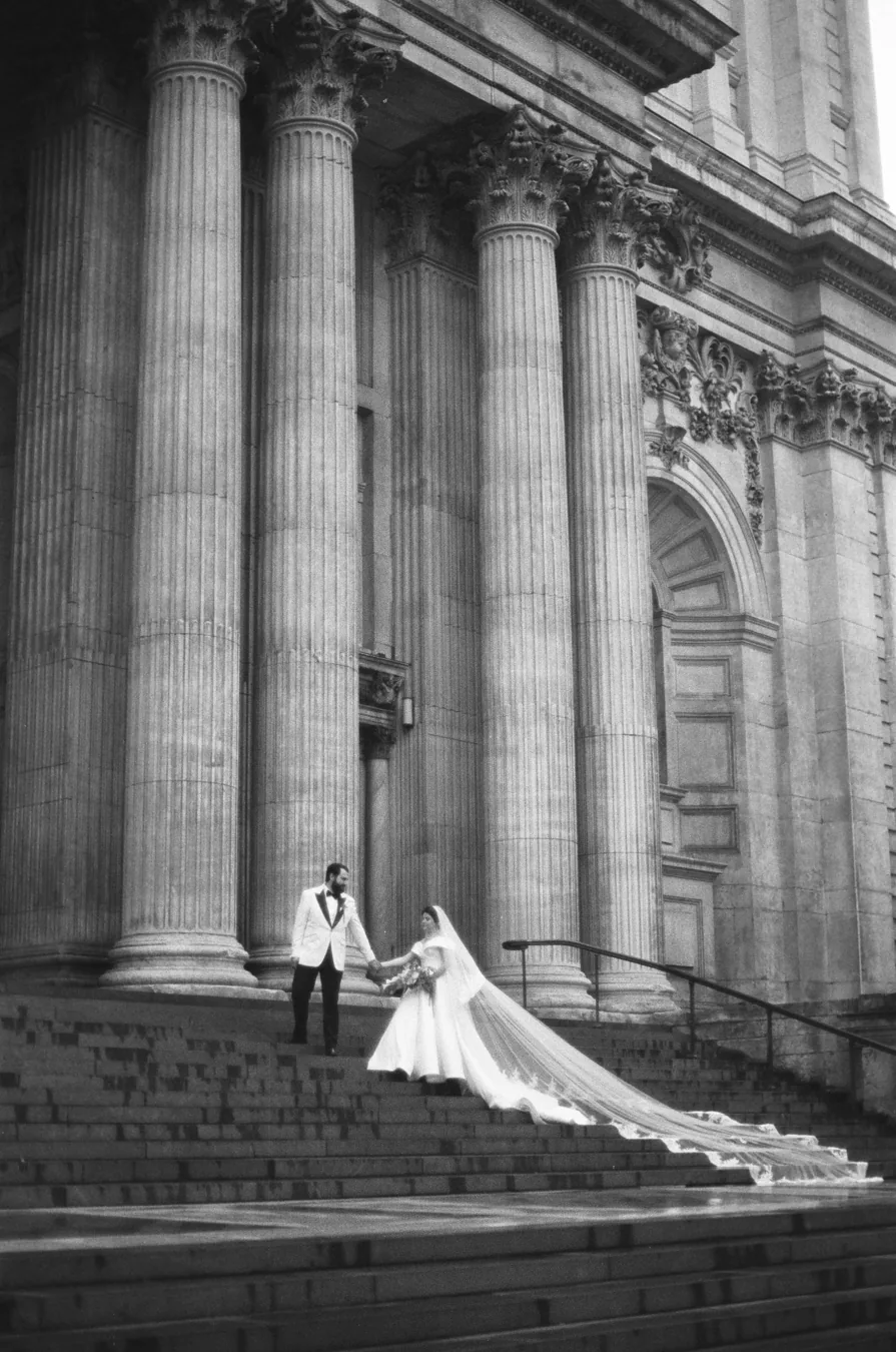 Black and white 35mm photograph of newlyweds holding hands on steps beneath stone columns