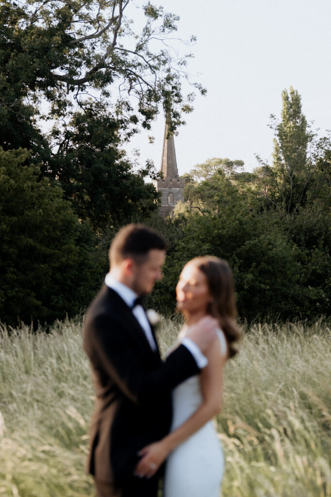Bride and groom embracing in foreground, church in focus in background